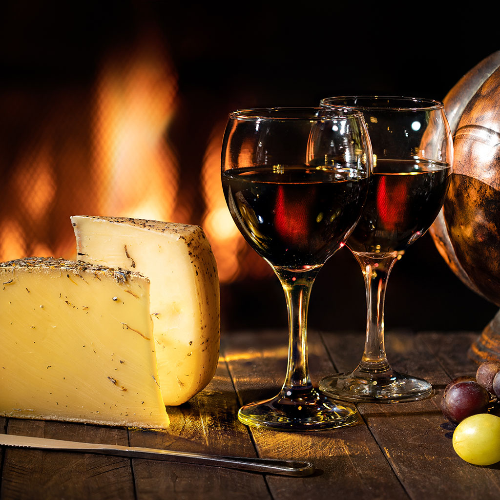 Wine cellar with wine glasses, cheese, grapes on rustic wooden table with fireplace in the background